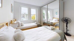 Une chambre d’hôtel avec deux lits simples, un grand miroir, un ventilateur sur pied et une fenêtre donnant sur des immeubles et des arbres.