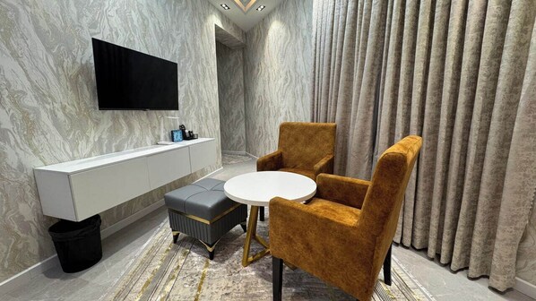 A modern room with a marble wall, a flat-screen TV, a white console, a round table, two velvet armchairs, and a patterned rug. - ShedevrPLAZA (Navoiy)