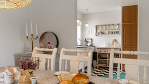 Un coin repas avec des chaises blanches, une table recouverte d’une nappe à motifs, un panier de pain et, au fond, une cuisine.