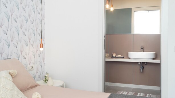 A bedroom with a bed, a nightstand, a lamp, and a bathroom with a sink and mirror. - 7Studios - 1 Sage (Milano)