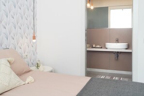 Een slaapkamer met een bed, een nachtkastje, een lamp en een badkamer met een wastafel en spiegel.