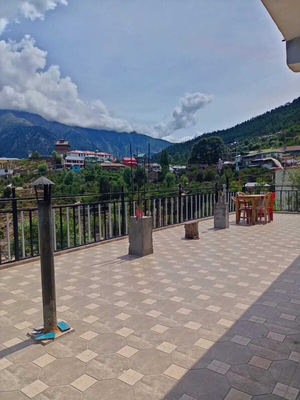 A terrace with a tiled floor, a railing, and a view of a mountainous landscape with buildings in the background. - Aarav Chaaras Homestay (Kalpa)