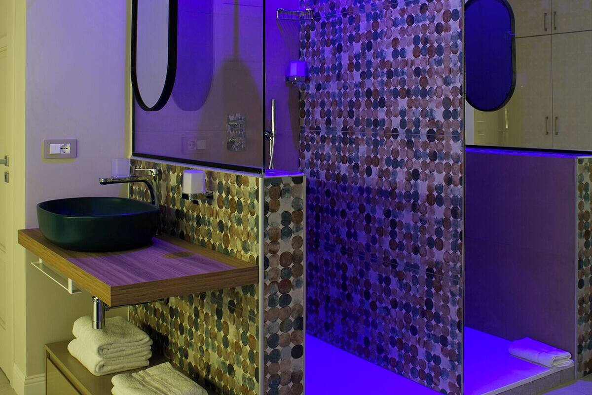 Ett modernt badrum med en stor spegel, ett grönt handfat och en duschhörna med en vägg som är klädd i mosaik.