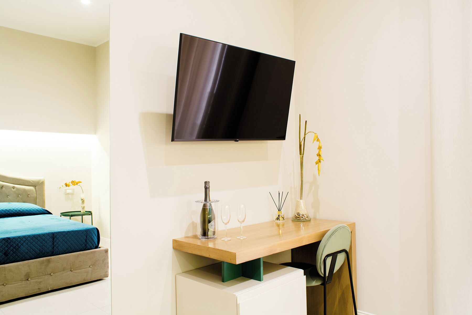 Une chambre d’hôtel moderne équipée d’un lit, d’un bureau avec une chaise, d’un téléviseur à écran plat fixé au mur et d’une petite table sur laquelle est posé un vase de fleurs.