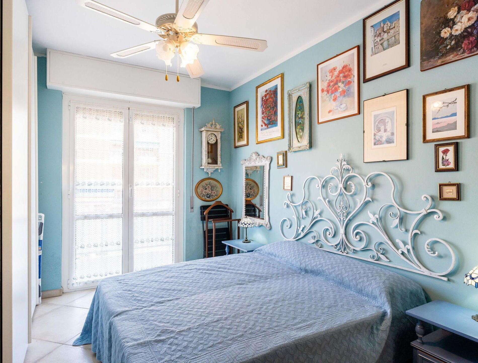 A bedroom with a bed, a ceiling fan, a window with lace curtains, and various framed pictures on the wall.