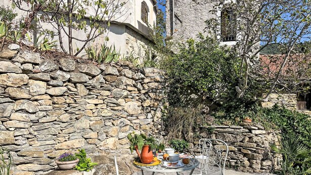 Un muro de piedra, un espacio al aire libre con sillas y una mesa metálicas, y un edificio con ventanas arqueadas.