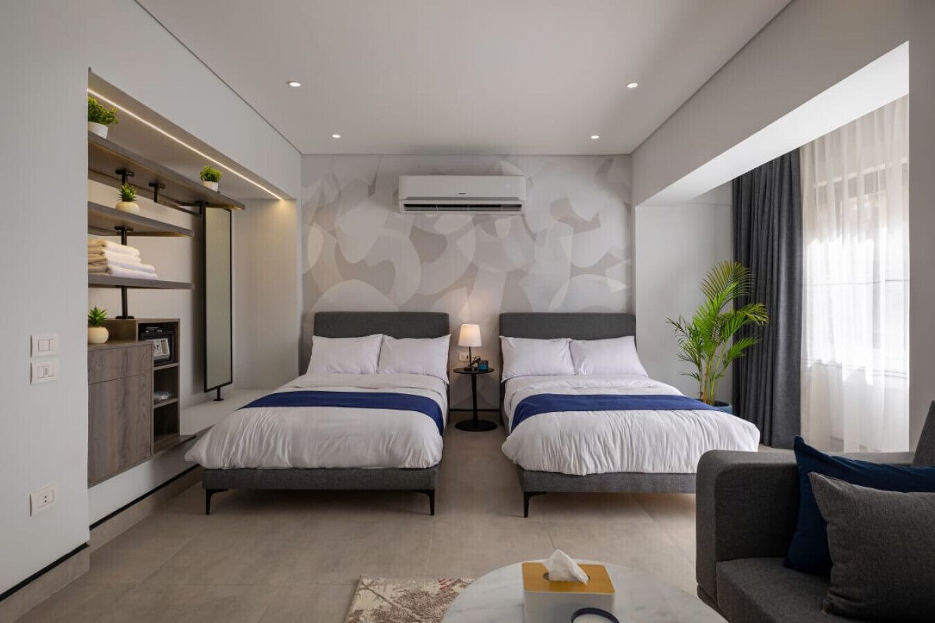 A modern hotel room with two beds, a sofa, a small table, a desk, a wardrobe, and a large window with curtains.
