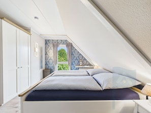 A bedroom with a sloped ceiling, a bed with patterned bedding, a window with curtains, and a view of greenery outside.
