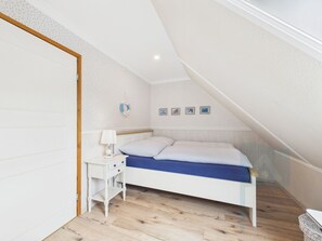Et soveværelse med skråvæg, en seng med blå madras, et sengebord med lampe og en væg med billeder i rammer.