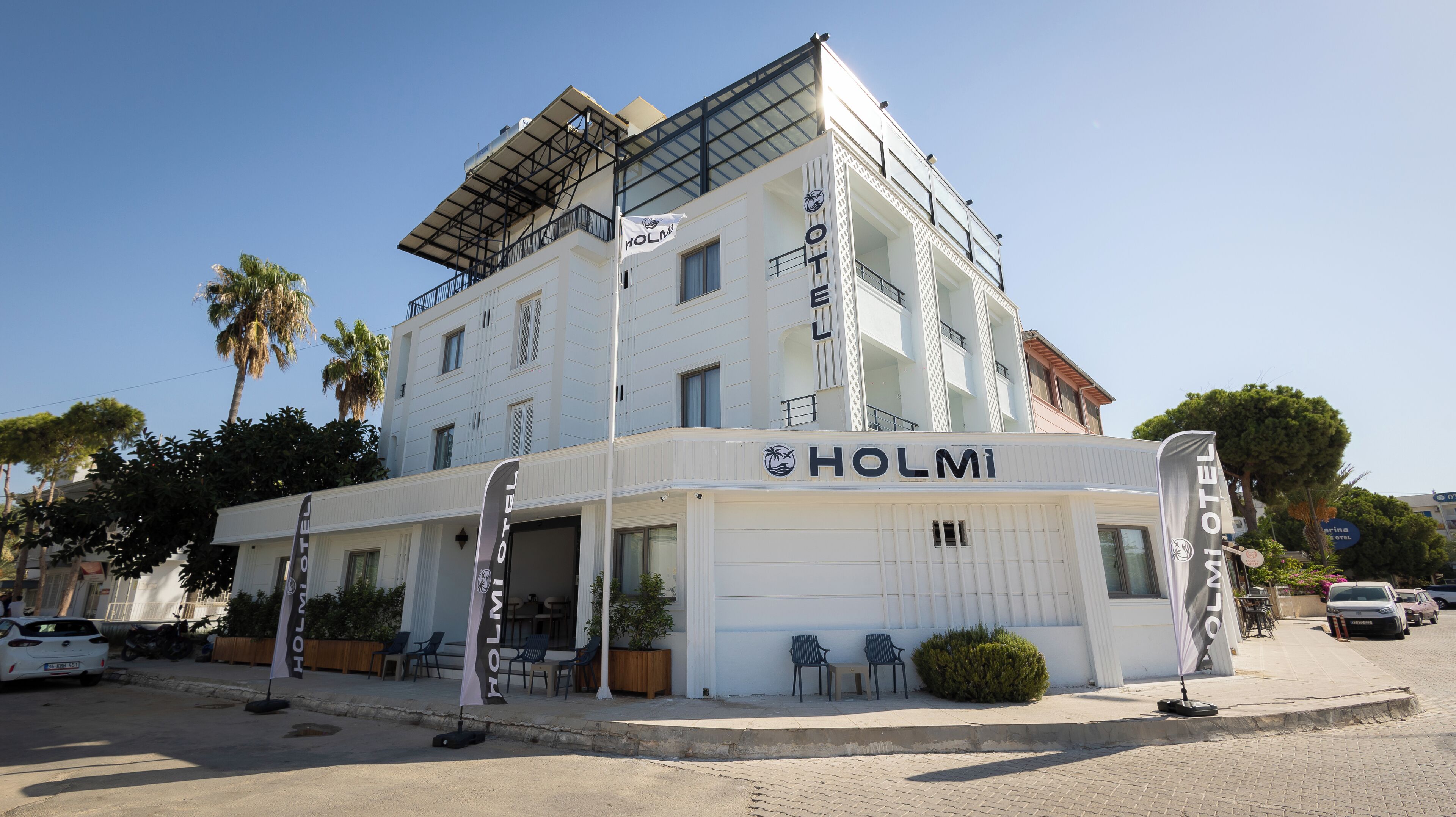 A white building with the name 'HOLMI HOTEL' on the facade, featuring a balcony and a covered outdoor seating area.