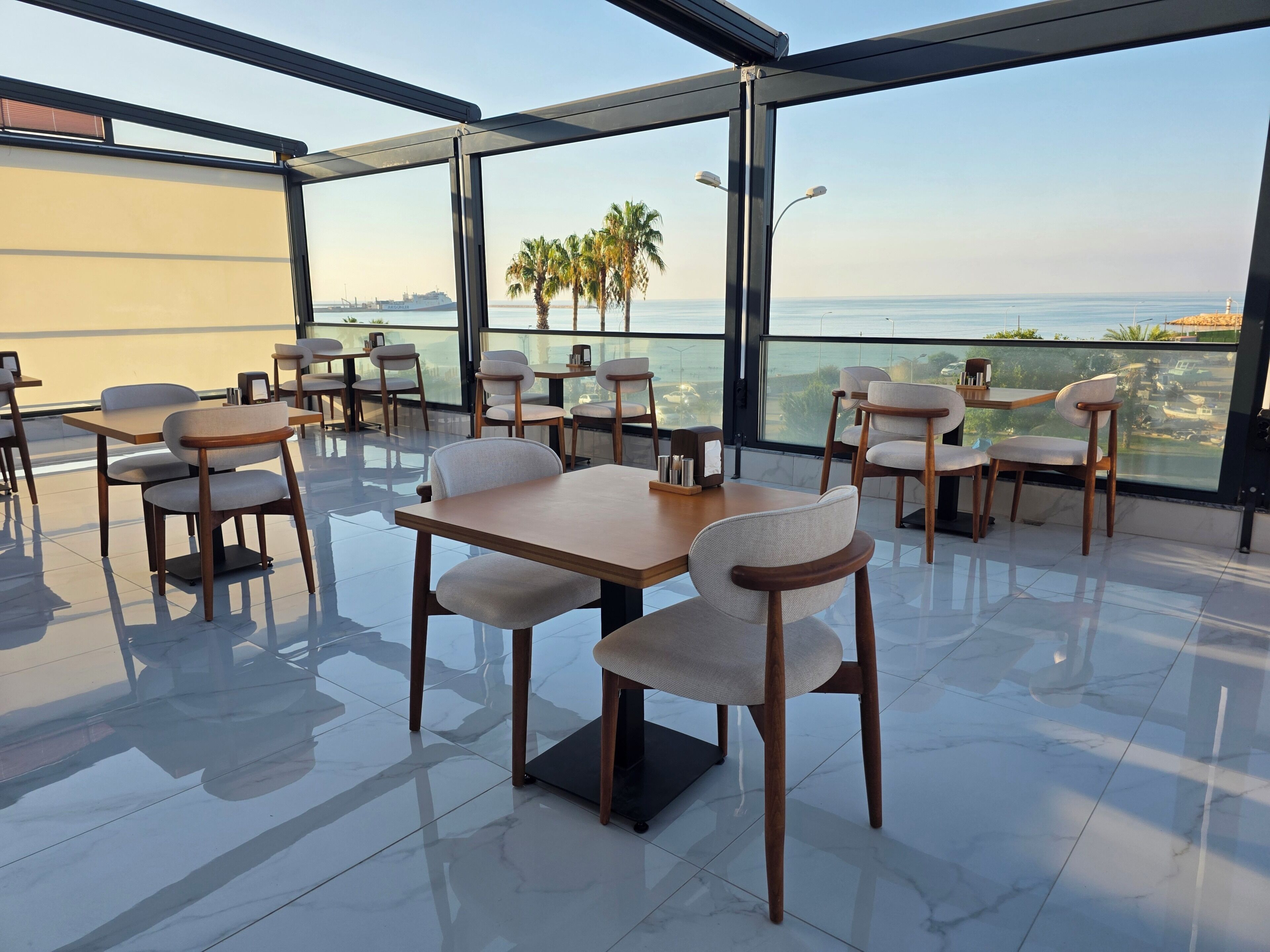 Um restaurante com mesas, cadeiras, janelas amplas e vista para o mar.