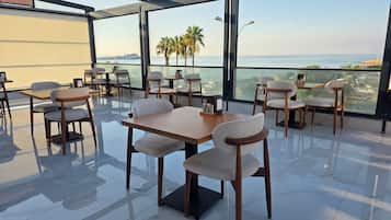 Um restaurante com mesas, cadeiras, janelas amplas e vista para o mar.