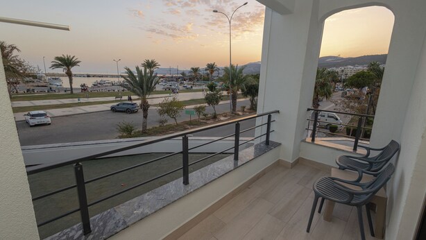 A balcony with a view of a marina, palm trees, and a road. - HOLMİ OTEL (Taşucu)