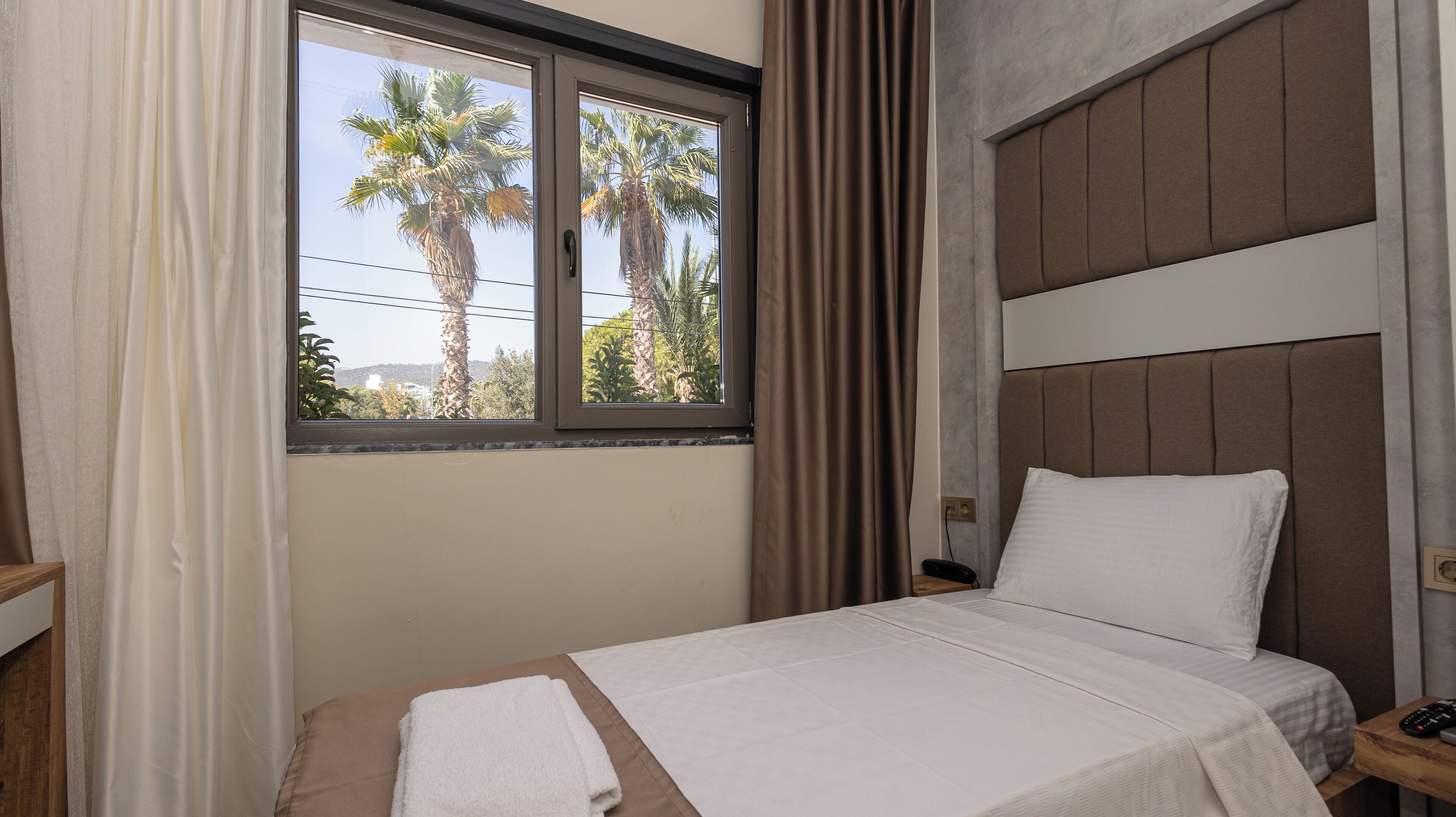 Una habitación de hotel con una cama, una mesita de noche, una ventana con cortinas y vistas a palmeras.