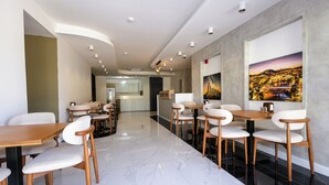 A modern hotel lobby with a marble floor, wooden tables and chairs, a reception desk, and a large cityscape painting on the wall. - HOLMİ OTEL (Taşucu)