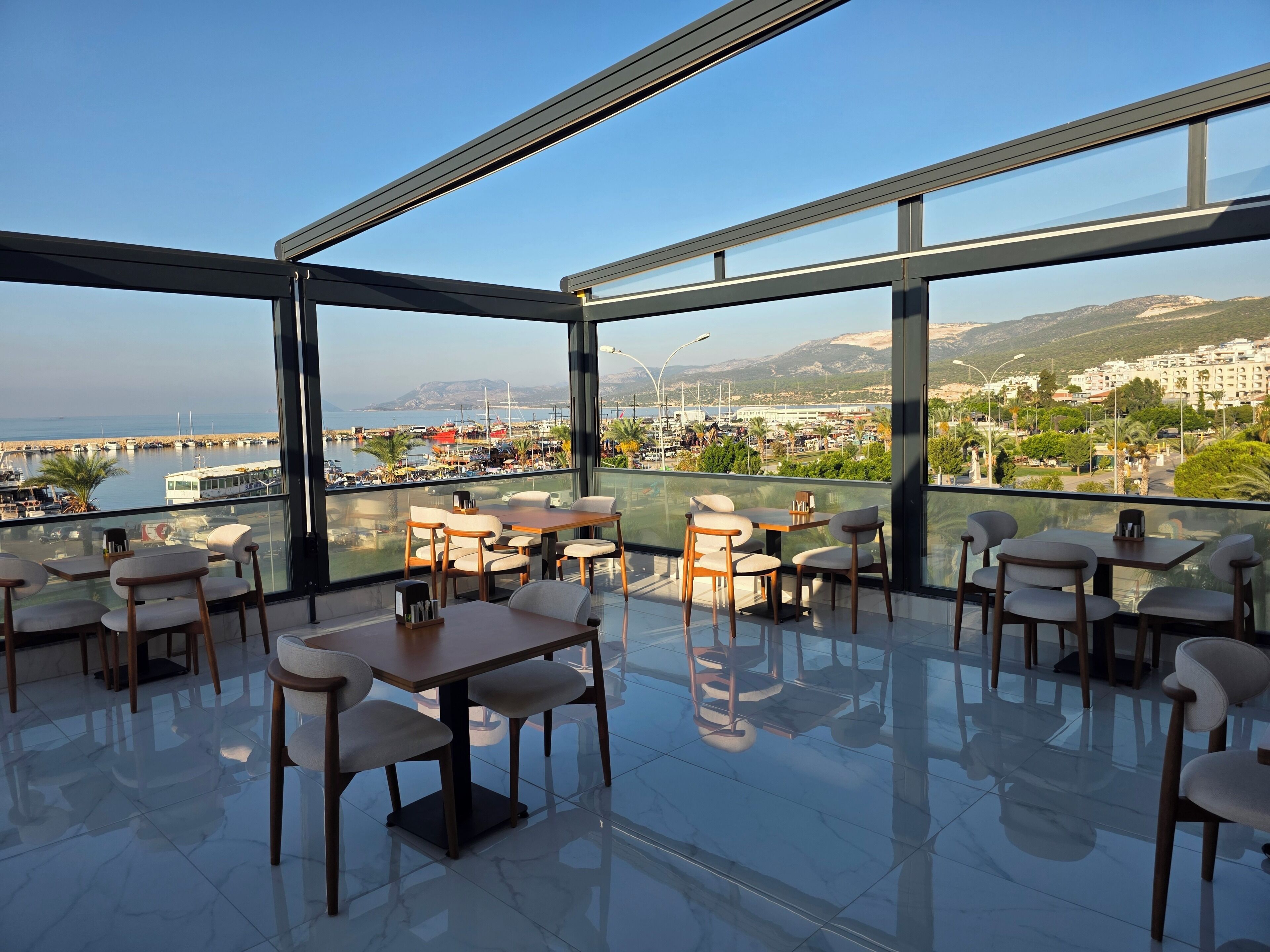 Un comedor moderno con paredes de vidrio, varias mesas y sillas, con vistas a un puerto deportivo y al horizonte urbano.