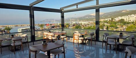 Un comedor moderno con paredes de vidrio, varias mesas y sillas, con vistas a un puerto deportivo y al horizonte urbano.
