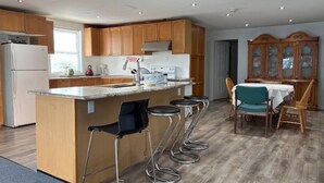 A modern kitchen with wooden cabinets, a central island with bar stools, a dining area with a table and chairs, and a large wooden cabinet with glass doors. - Beautiful Riverfront House (Campbellford)