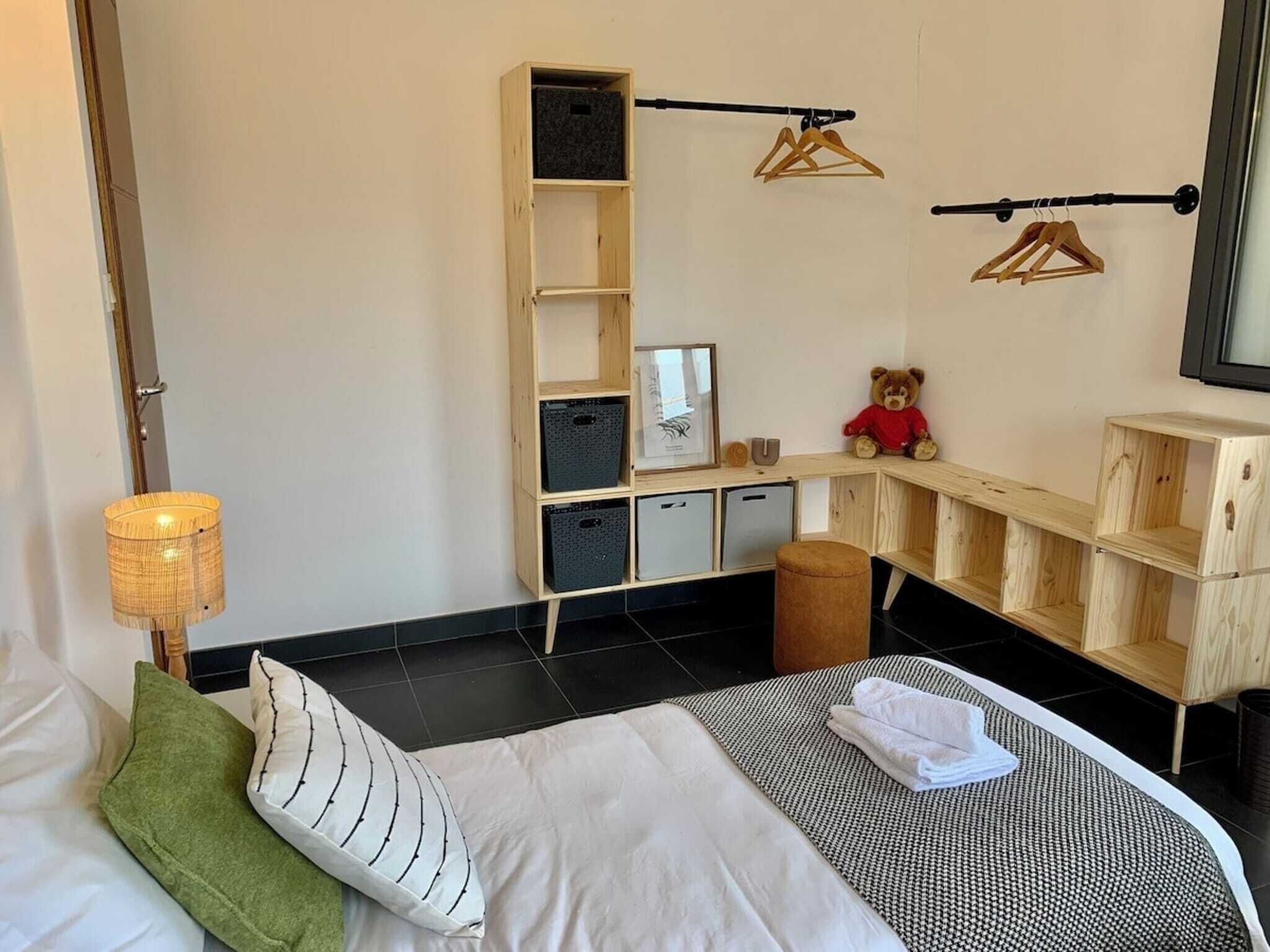 A hotel room with a bed, a wooden wardrobe, a small stool, a teddy bear, and a hanging clothes rack.