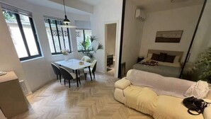 A modern living room with a herringbone floor, a sofa, a dining table with chairs, and a bed with a headboard.