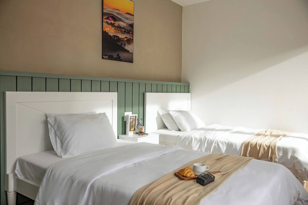 Una camera d'albergo con due letti, un comodino con una lampada, un quadro appeso al muro e un vassoio per la colazione sul letto.