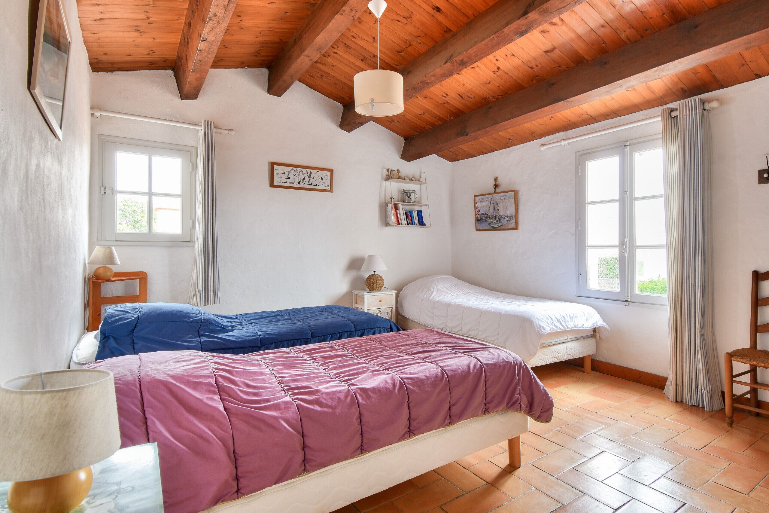 Een slaapkamer met twee bedden, houten balken in het plafond, een raam en een plank met boeken.