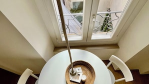 Een ronde tafel met een gevlochten mand als middelpunt, twee witte stoelen en een raam met uitzicht op een gebouw en groen.
