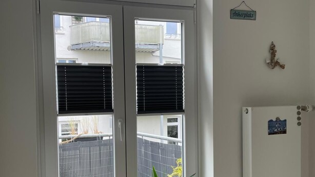 Una habitación con puerta de vidrio corrediza que da a un balcón, una planta en maceta en el alféizar de la ventana, un refrigerador blanco y una pequeña señal que dice 'Ankerplatz'.