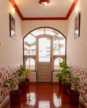 Un pasillo con una puerta blanca, flanqueado por dos ventanas que dan a vistas de palmeras. Hay plantas en macetas a cada lado de la puerta.