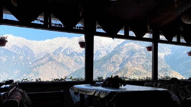 Una habitación con una mesa, bancos, ventanas amplias y vistas a la montaña, y plantas colgantes.