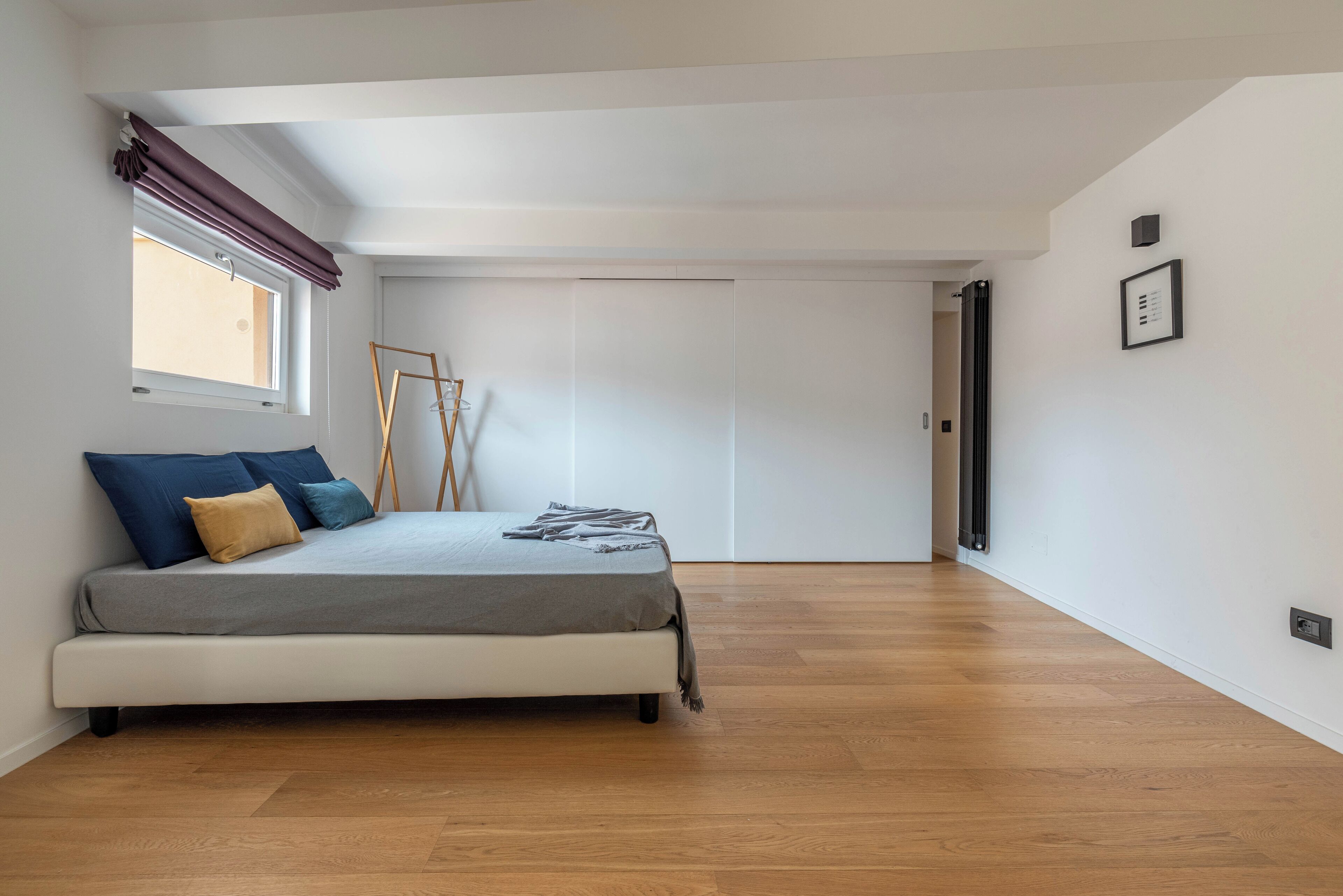 Ein Schlafzimmer mit einem Bett, Holzboden, einem fenster mit Vorhängen, einer Tür und einem an der Wand befestigten Lichtschalter.