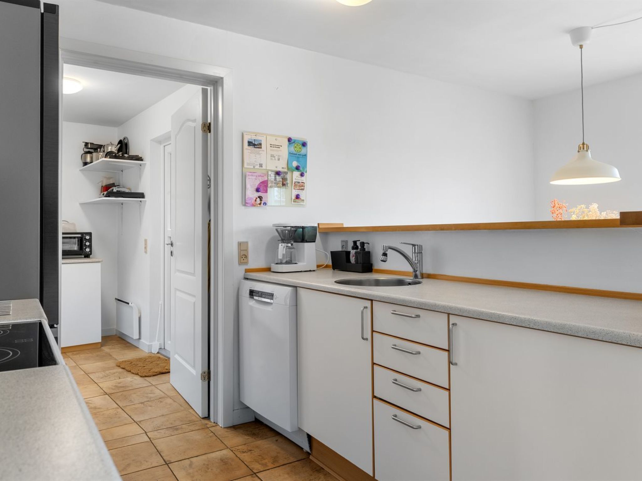 A compact kitchen with a built-in dishwasher, a coffee maker, and a sink. There is a door leading to another room, and a wall-mounted bulletin board with various papers.