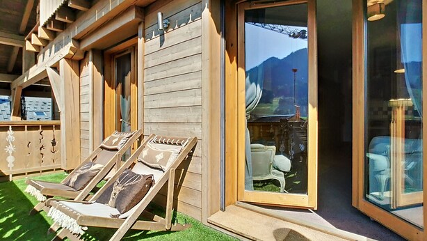 Un edificio de madera con grandes ventanales y vistas a las montañas. Hay dos sillones con cojines en el exterior.