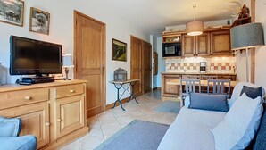 Una sala de estar con televisor, muebles de madera y una cocina con alacenas de madera y un microondas.