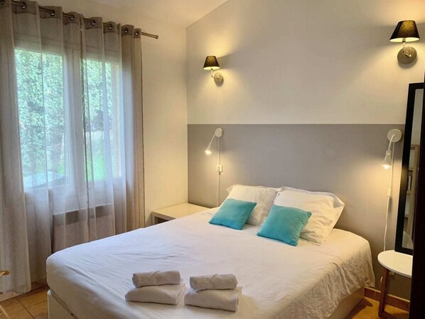 A neatly made bed with white linens and two blue pillows, a bedside table with a lamp, and a window with sheer curtains.