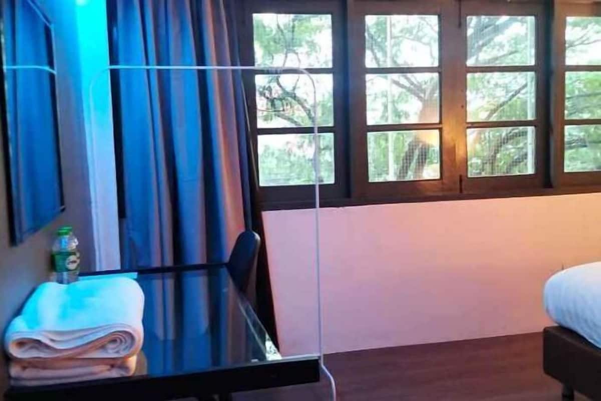 A hotel room with a bed, a window with curtains, a table with a folded towel and a bottle, and a wall-mounted TV.