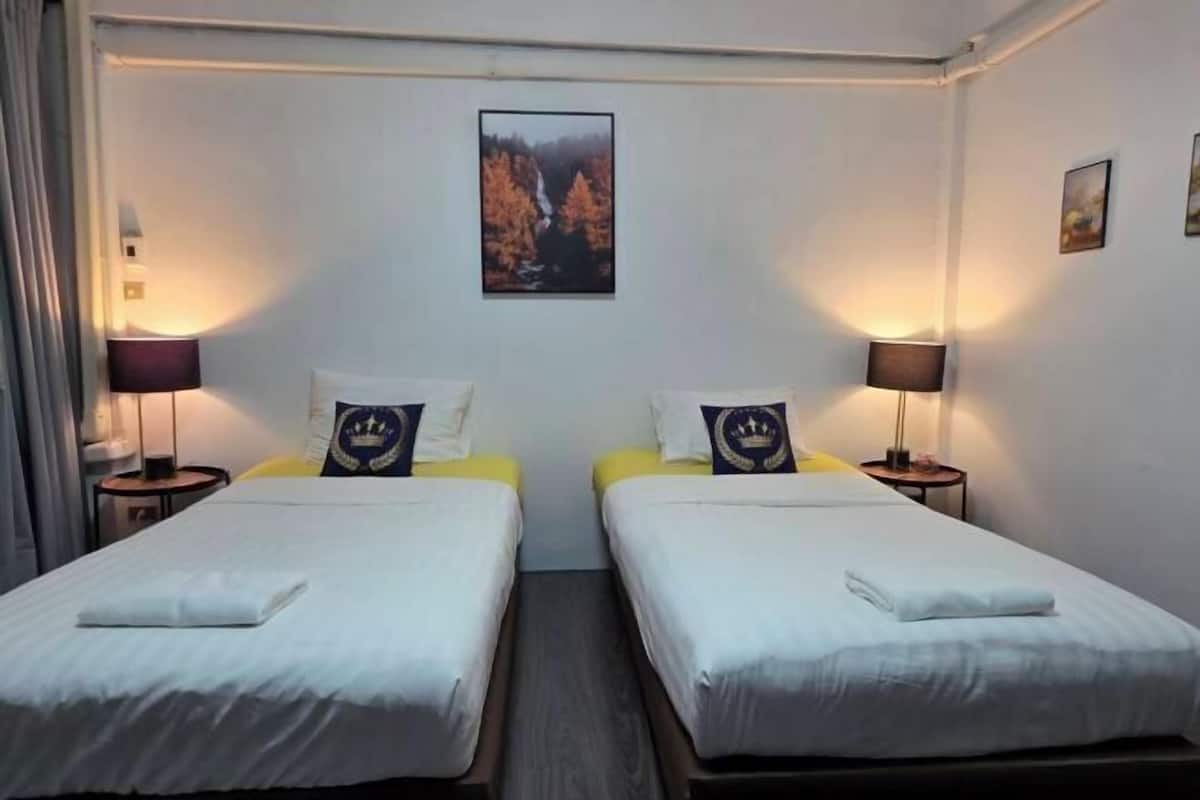 A hotel room with two single beds, each with a pillow and a small table. There is a framed picture on the wall above the beds.