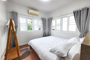 Une chambre à coucher avec un lit, des fenêtres, des rideaux, un appareil de climatisation et une chaise en bois.