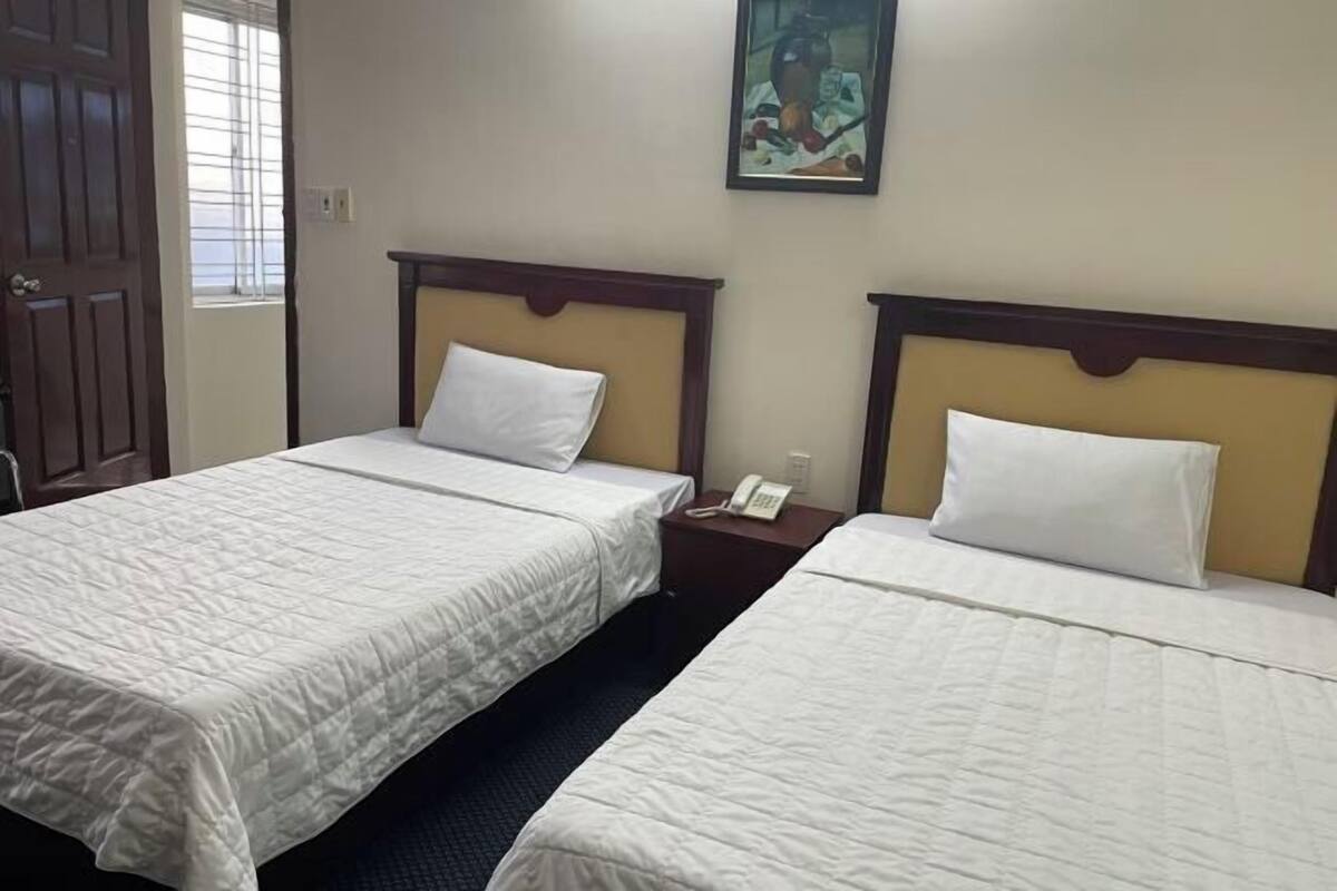 Une chambre d’hôtel avec deux lits, chacun doté d’une tête de lit en bois et de draps blancs. Un tableau est accroché au mur au-dessus des lits, et un téléphone se trouve sur la table de chevet.