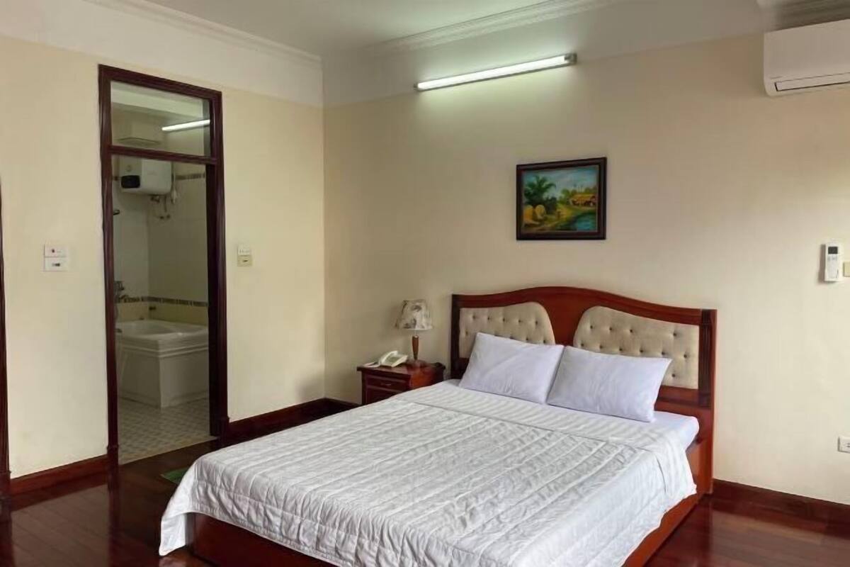 Une chambre d’hôtel avec un lit, une table de chevet, une lampe, un tableau au mur et une salle de bain visible à travers une porte ouverte.