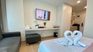 Habitación de hotel con cama, sofá, televisión montada en la pared, una mesita con un jarrón de flores y una cocina visible a través de una puerta abierta.