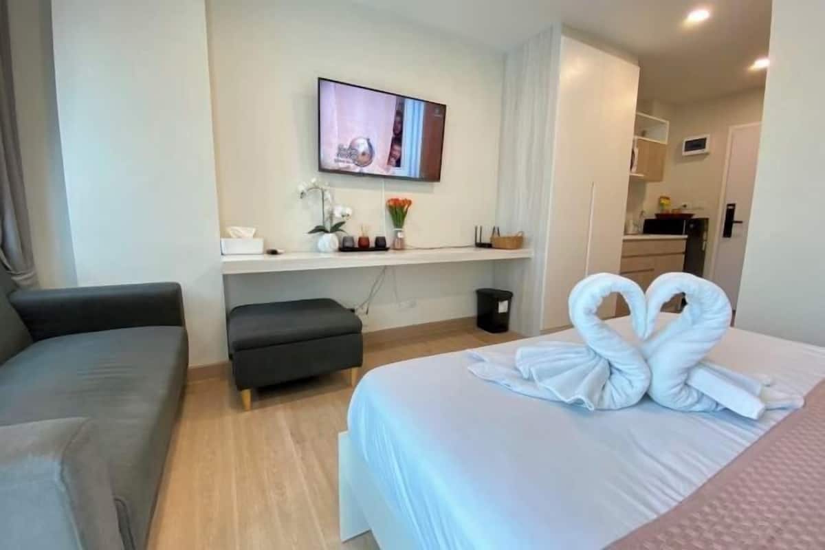A hotel room with a bed, a sofa, a TV mounted on the wall, a small table with a vase of flowers, and a kitchen area visible through an open door.