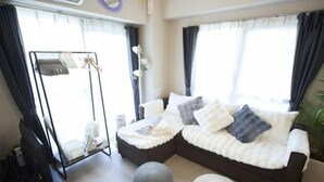 A modern living room with a sectional sofa, a standing TV, a small round ottoman, and a clothing rack. - Mezon Etowaru #706 (Nagoya)