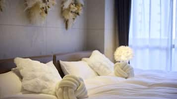 Cama com roupa de cama branca e almofadas decorativas, luminária fixada na parede e janela com cortinas.