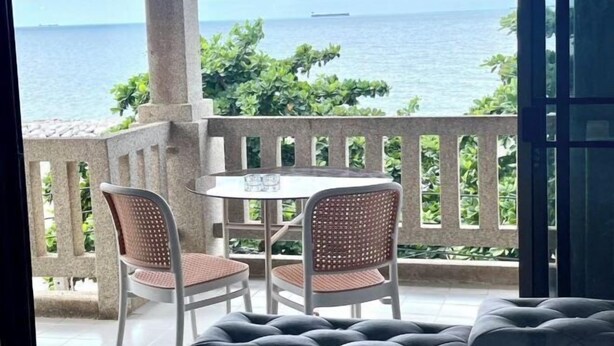 Un balcone con due poltrone in vimini e un tavolino, affacciato su uno specchio d'acqua con una nave all'orizzonte.