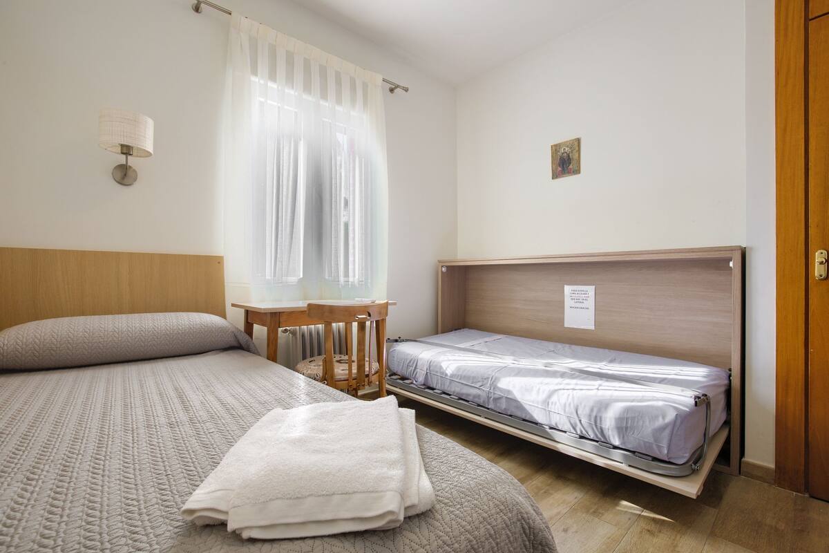 Une chambre d’hôtel avec deux lits, une tête de lit en bois, une petite table et une fenêtre avec des rideaux légers.