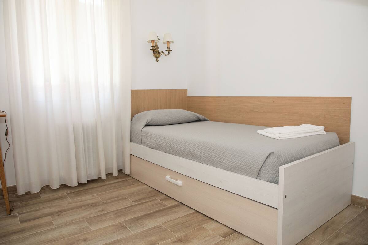 Un lit simple avec une tête de lit en bois, une table de chevet blanche et un luminaire fixé au mur.