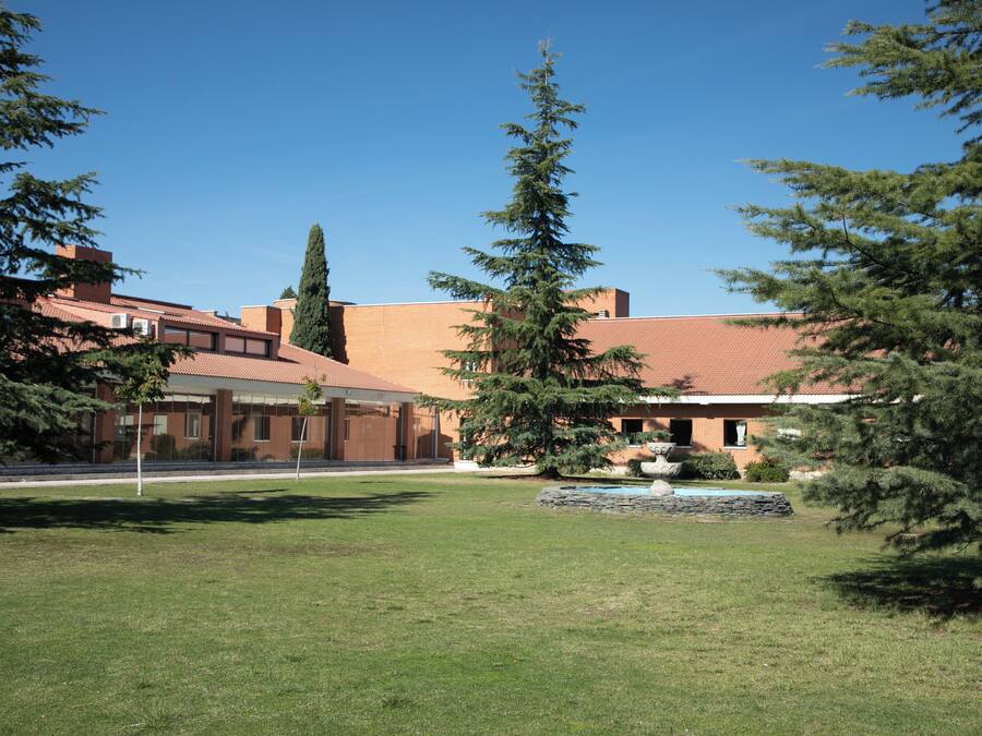 A building with a brick facade and a tiled roof, surrounded by a well-maintained lawn and several evergreen trees.