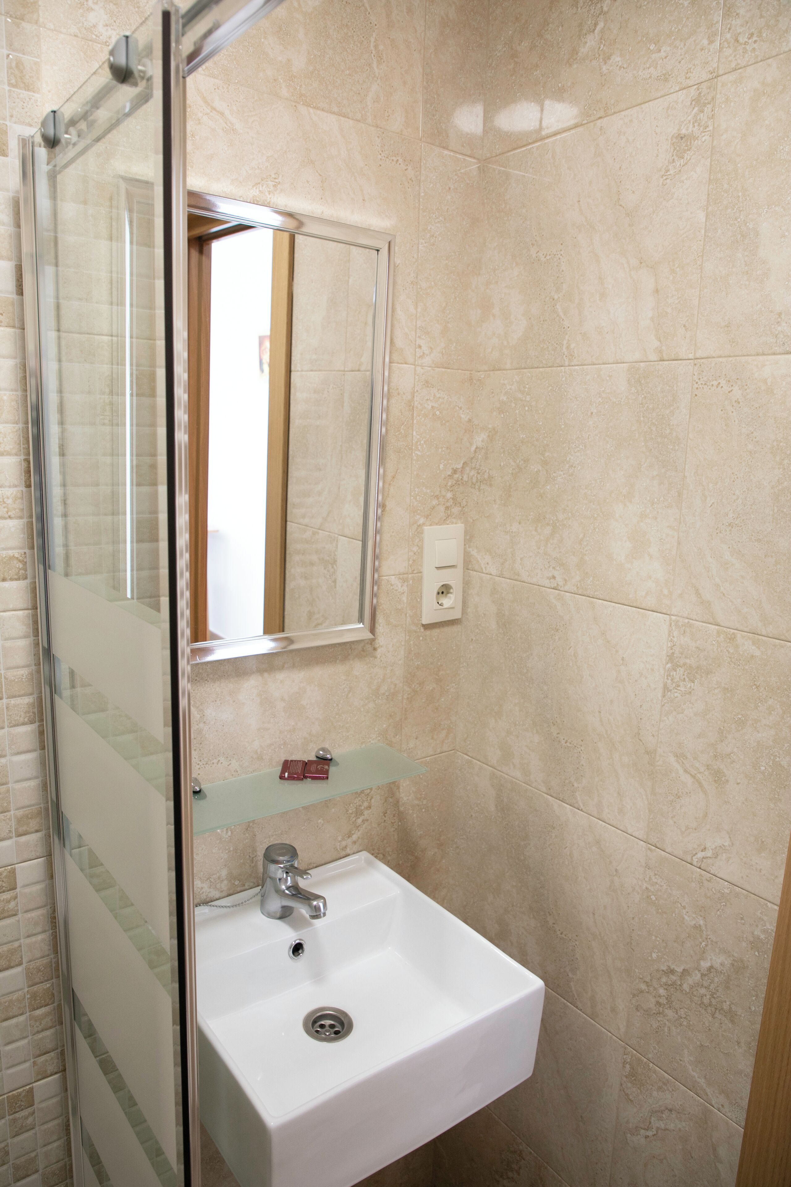 A bathroom with a glass shower enclosure, a white sink with a mirror above it, and beige tiled walls.
