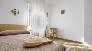 Un letto rifatto con un copriletto beige, un piccolo tavolo in legno con una lampada e una sedia.